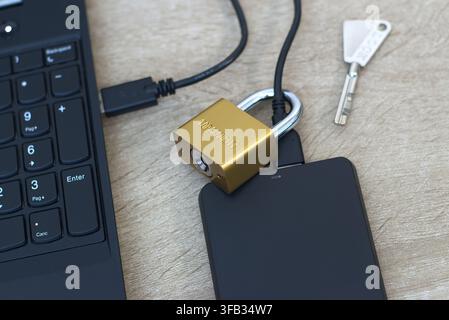 Close-up view of a small brass padlock physically secured around the USB cable of a black external hard drive. Laptop keyboard visible on the side. Co Stock Photo