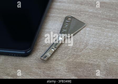 Detailed view of a metal security key resting on a wooden background, with a blurred smartphone in the background. Illustrates the theme of physical t Stock Photo