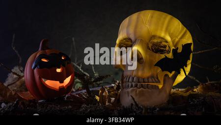 Creepy glowing human skull with a bat on it and smiling pumpkin lantern placed among withered leaves and branches on a dark background. Stock Photo