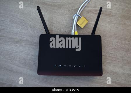 A gold padlock locking network ethernet cables connected to a wireless router on a wooden desk. A conceptual image illustrating physical cybersecurity Stock Photo
