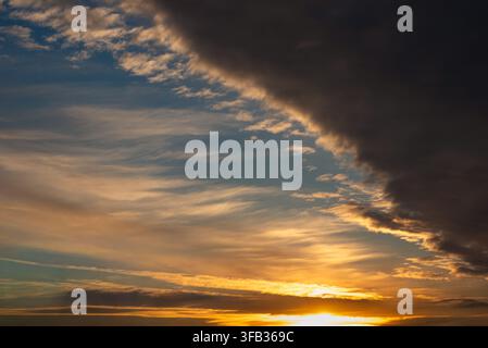 Beautiful sunset landscape featuring vibrant golden light at the horizon, wispy cirrus clouds in a blue sky, and a large dramatic dark cloud formation Stock Photo
