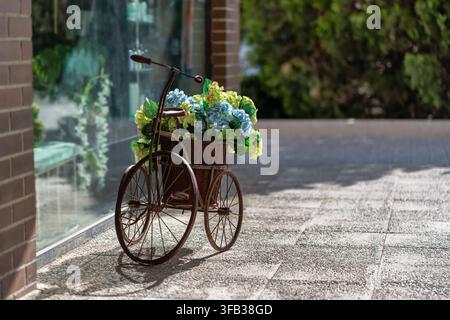 Decorative tricycle with a wicker basket of colorful flowers outside a sunlit shop window Stock Photo