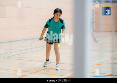 Female badminton player prepares to serve during an indoor tournament match, holding a racket and shuttlecock on a marked sports court with a competit Stock Photo
