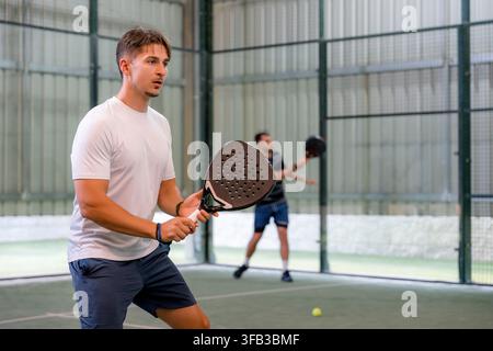 Focused male player holds a padel racket in ready position on an indoor court while an opponent prepares to return the ball during a competitive tourn Stock Photo
