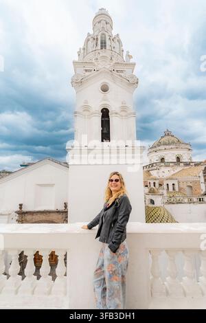 Smiling woman in sunglasses stands on the rooftop of the Church of the Sanctuary in Quito, Ecuador, framed by ornate white towers, tiled domes, and dr Stock Photo