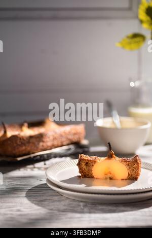 Close-up of a slice of pear sponge cake topped with poached pear on a white plate, with the remaining cake, bowl, spoon, and yellow flowers in warm su Stock Photo
