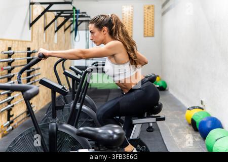 Athletic woman in sportswear exercises on an air bike at a modern gym, gripping the moving handles during an intense cardio workout with fitness equip Stock Photo
