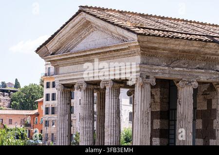 Sunlit view of an ancient Roman temple in Rome, Italy, featuring weathered stone columns, a triangular pediment, tiled roof, and historic urban buildi Stock Photo