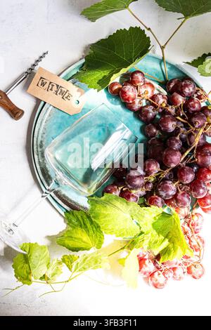 Overhead still life of red grape clusters with green vine leaves, an empty wine glass, corkscrew and wine tag arranged on a turquoise ceramic plate. Stock Photo