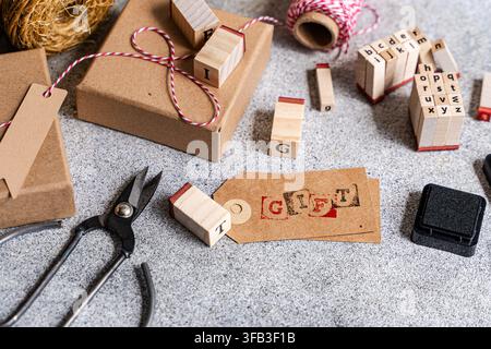 Overhead view of handmade gift wrapping materials with kraft paper boxes, alphabet stamps, red twine, blank tags, ink pad and scissors on a gray textu Stock Photo