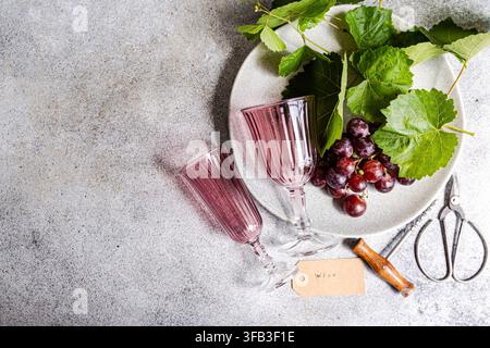 Overhead still life of pink wine glasses, fresh red grape clusters and green leaves on a plate, arranged with garden shears, a cork and blank tag on t Stock Photo