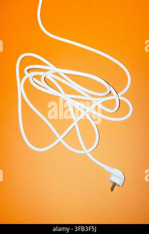White electrical power cord with a two-pin plug tangled across a vibrant orange background, viewed from above. Minimal studio composition for electric Stock Photo