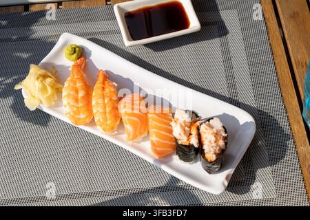 Close-up of assorted nigiri sushi with salmon, shrimp, white fish, avocado, and pickled ginger arranged on a white ceramic platter over a woven table Stock Photo