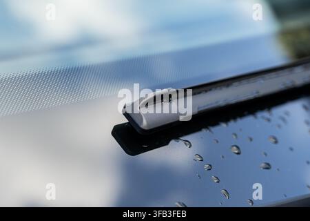 Close-up of an electric car windshield wiper resting on wet glass, with water droplets and soft blue reflections highlighting vehicle maintenance, wea Stock Photo