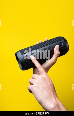 Close-up of a woman's hand holding and pressing controls on a black portable wireless speaker against a vivid yellow background, showcasing modern aud Stock Photo