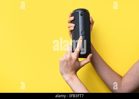 Close-up of a woman’s hands holding and operating a black portable Bluetooth speaker against a bright yellow background, showcasing wireless audio tec Stock Photo