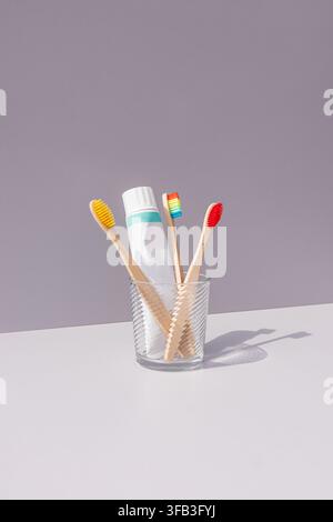 Three bamboo toothbrushes with colorful bristles and a toothpaste tube stand in a clear glass cup on a white surface and soft gray background. Stock Photo