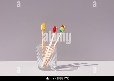 Three bamboo toothbrushes with yellow, red, and rainbow bristles stand in a textured glass cup on a white surface, casting long shadows against a gray Stock Photo