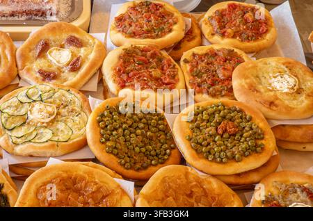 A selection of round mini pizzas is arranged on a table at a food market. These pizzas feature toppings like red peppers, green peas, caramelized onio Stock Photo
