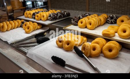Fresh golden donuts and chocolate glazed donuts arranged on serving trays at a breakfast buffet with serving tongs. Stock Photo