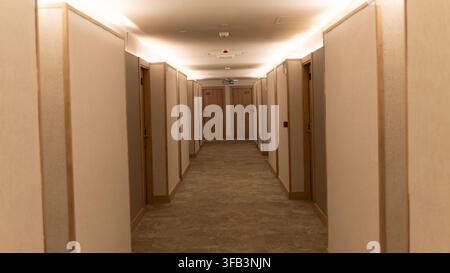 Long hotel corridor with numbered guest room doors, warm lighting, carpeted floor and an emergency exit sign at the end. Stock Photo