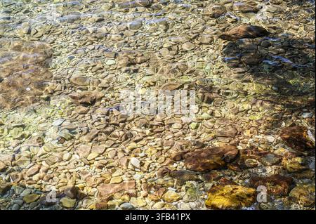 Sunlight dances on a clear riverbed with smooth, colorful stones and pebbles. Stock Photo