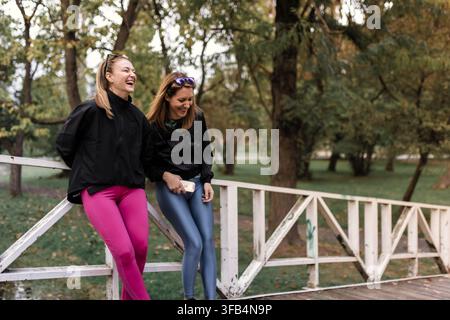 Friends laugh together on a bridge in a park during a sunny afternoon in fall while enjoying each others company and taking a break from their Stock Photo