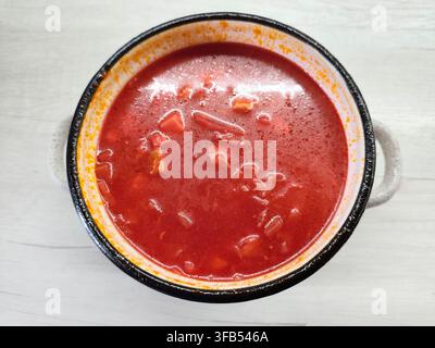Red borscht soup with cabbage and meat served in a small pot with a metal spoon on a light wooden table surface, top view photo. Stock Photo