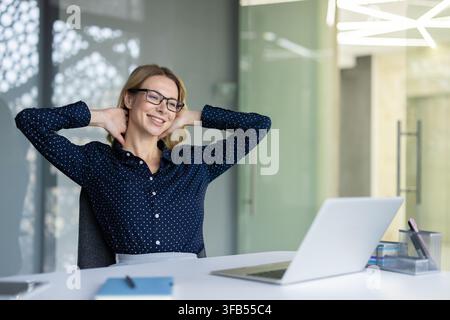 Happy young businesswoman in glasses stretching arms behind head, smiling confidently while sitting at her office desk, taking a well-deserved break after successful project completion Stock Photo