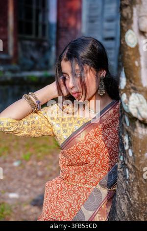 A woman in a traditional patterned saree stands leaning against a tree trunk in an outdoor setting with a closed shuttered building in the background. Stock Photo