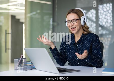 Businesswoman with glasses and wireless headset smiling during a virtual meeting on her laptop in a modern office, participating in remote work, webinar or online coaching session Stock Photo
