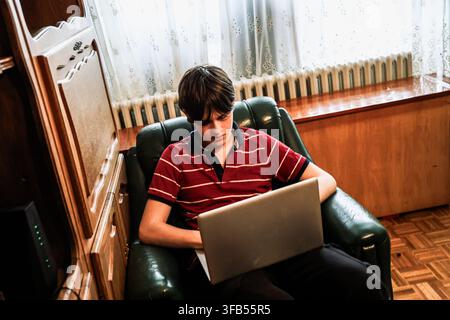 A boy sits on a green chair in a simple living room. He uses a laptop with focus. Sunlight comes through the window and falls on the wooden floor. Stock Photo
