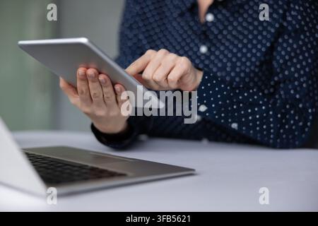 Person is using a digital tablet, holding the device with both hands while tapping the screen, with a laptop partially visible on a white desk, representing modern work and connectivity Stock Photo