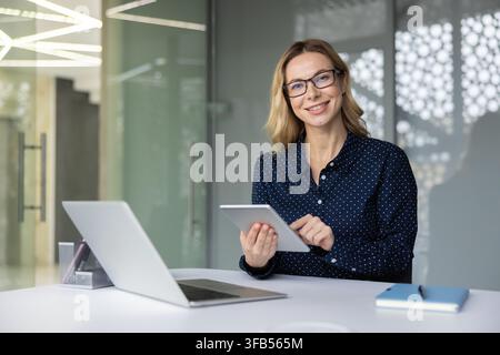 Confident businesswoman smiling at camera while working at her modern office desk with laptop and tablet, managing projects and communicating digitally in a bright corporate workspace Stock Photo