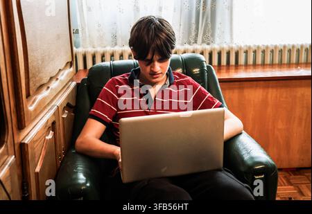 A boy sits in a green chair focused on his laptop. Sunlight streams through a window. The room has wooden features and simple decor. Stock Photo
