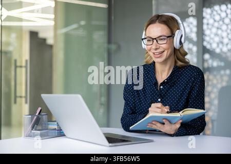 Woman in headphones and glasses smiling while writing notes in a notebook, actively participating in a virtual meeting or remote education session at her office desk Stock Photo