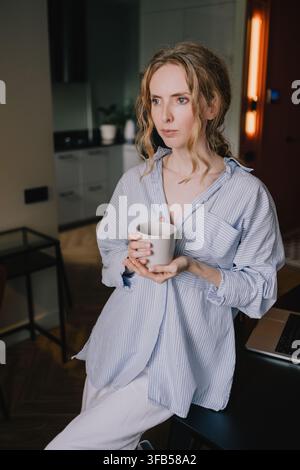 Woman holding a cup during a quiet moment beside a laptop at home, reflecting a morning routine, remote work lifestyle and work-life balance Stock Photo