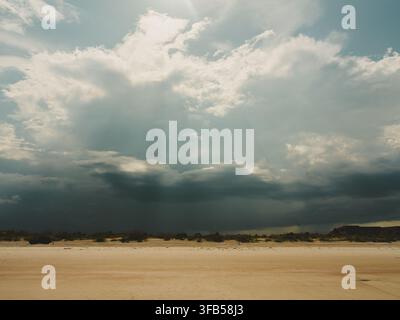 Dramatic storm clouds and shafts of sunlight over a wide sandy beach and distant coastal horizon. Stock Photo