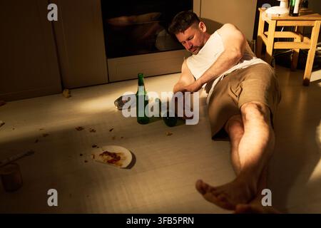 Depressed Man Lying on Kitchen Floor with Alcohol Bottles Stock Photo