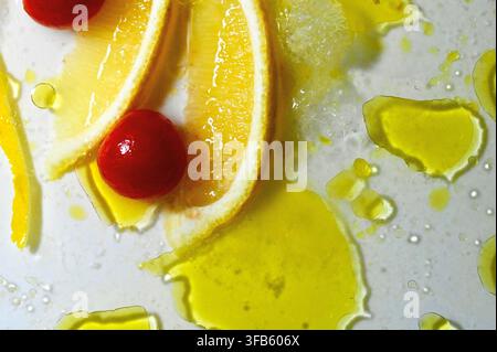 A macro photograph of fresh lemon slices, two whole cherry tomatoes, and lemon zest arranged with glistening drops and pools of extra virgin olive oil and small oregano leaves on a white surface. Stock Photo