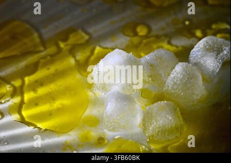 A horizontal studio macro photograph of melting ice cubes surrounded by pools and glistening drops of extra virgin olive oil on a white surface. Stock Photo