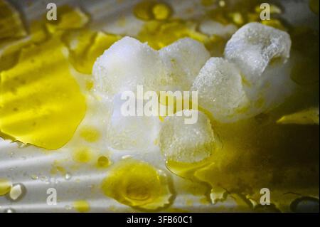 A horizontal studio macro photograph of melting ice cubes surrounded by pools and glistening drops of extra virgin olive oil on a white surface. Stock Photo