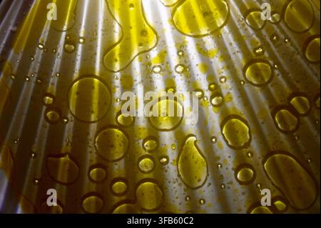 A horizontal macro photograph featuring scattered golden drops and pools of olive oil on a white ribbed surface, illuminated by dramatic light rays creating bright highlights and deep shadows. Stock Photo