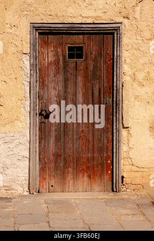 Weathered wooden door with small square window set in sunlit rough plaster wall, framed by aged stone trim, rusted hardware Stock Photo