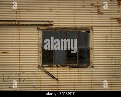 A weathered, rusty corrugated metal wall with an old multi-pane glass window of an abandoned industrial building Stock Photo
