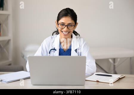 Smiling South Asian female doctor using laptop in clinic office Stock Photo