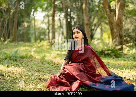 A woman sits cross-legged on the ground wearing a red saree with a dark blue border in a wooded area with sunlight filtering through the trees. Stock Photo
