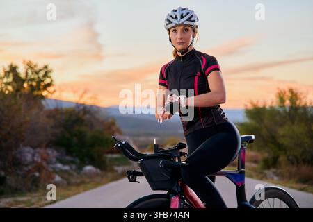 Female Cyclist Checking Watch on Road Bike During Sunset Training Ride in Rural Landscape Stock Photo