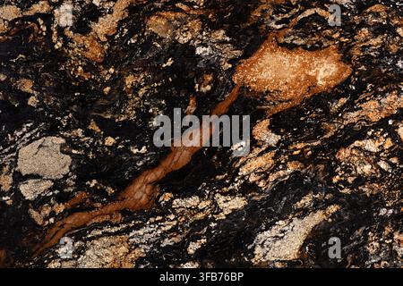 Detail of a natural granite slab showing dark background with copper orange veins and speckled golden mineral inclusions on a glossy polished surface. Stock Photo