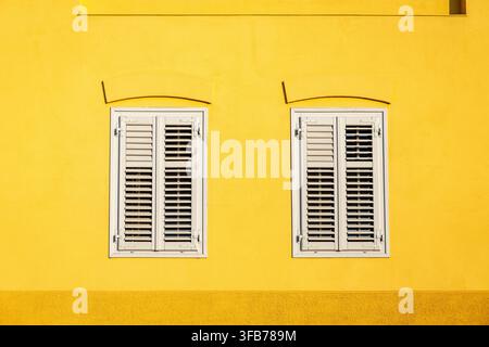 Two White Shuttered Windows on Bright Yellow Wall. Two white shuttered windows set in a vivid yellow stucco wall, creating a bright symmetrical Stock Photo